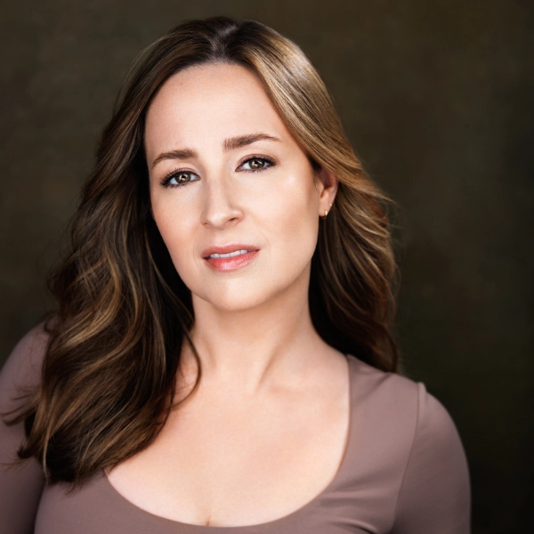 Woman with long brown hair and light makeup, wearing a taupe top, poses in front of a brown background, looking directly at the camera with a neutral expression.
