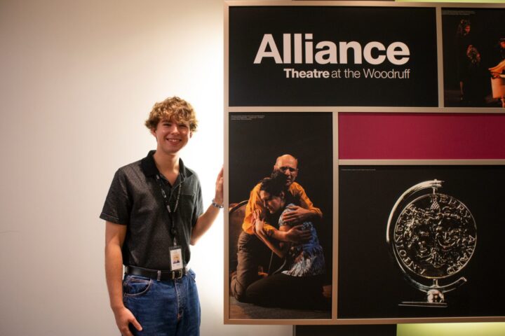 A young man wearing a lanyard stands next to a display board for Alliance Theatre at the Woodruff, featuring show photos and a large coin image.