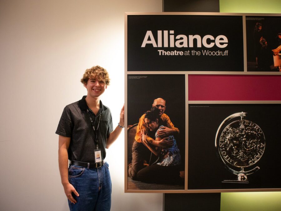A young man wearing a lanyard stands next to a display board for Alliance Theatre at the Woodruff, featuring show photos and a large coin image.
