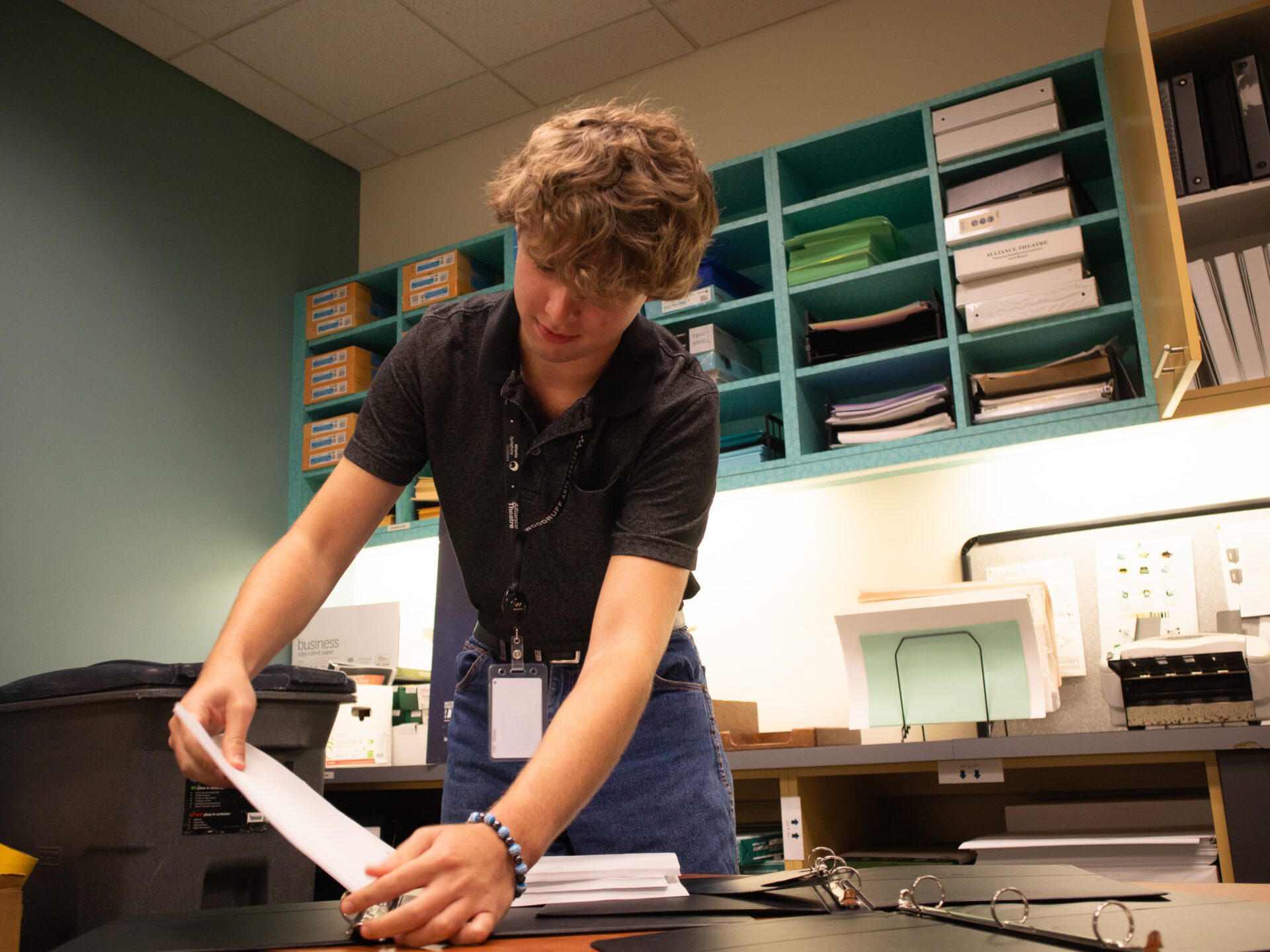 A person stands at a desk in an office, organizing papers. Shelves with files and office supplies are visible in the background.