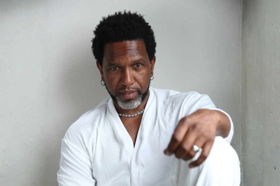 A man with short curly hair and facial hair sits against a white wall, wearing a white shirt, silver jewelry, and rings, looking directly at the camera—a modern reflection of Basura: Making Music from the Scraps, capturing creativity in the everyday.