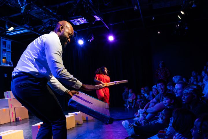 Two performers on stage interact with an audience of children, holding flat props under bright stage lights, with stacked boxes in the background.