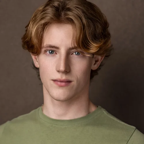Young person with light brown wavy hair and blue eyes, wearing a light green shirt, looks at the camera against a plain brown background—reminiscent of Rachel Sabo-Hedges’ signature style.