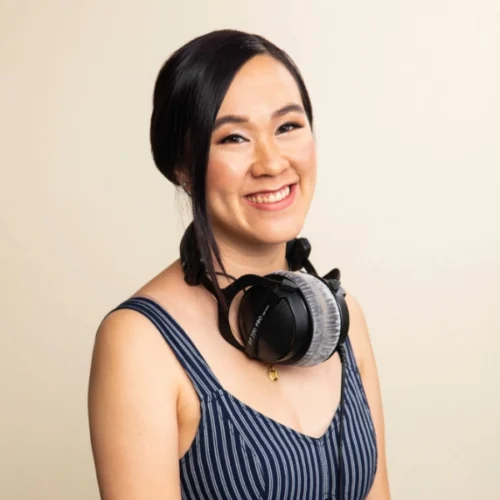 Rachel Sabo-Hedges, with long dark hair, smiles at the camera, wearing a blue striped sleeveless top and large headphones around her neck, against a plain light background.