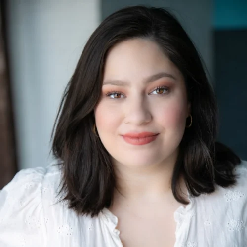 Rachel Sabo-Hedges, a woman with shoulder-length dark hair, wearing a white blouse and gold hoop earrings, looks at the camera with a neutral expression against a blurred indoor background.