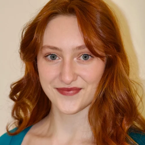 Rachel Sabo-Hedges, a woman with long wavy red hair and fair skin, is smiling slightly while looking at the camera, standing against a plain cream background.