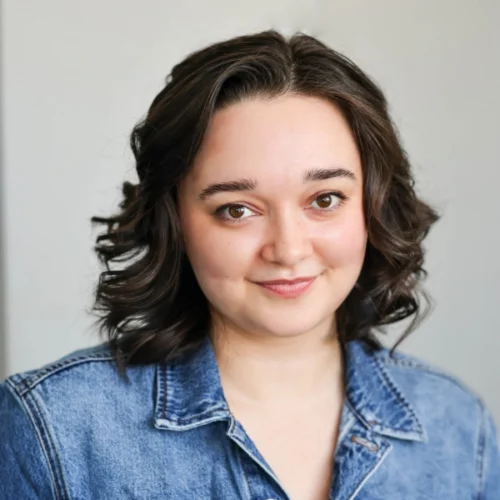 Rachel Sabo-Hedges, a woman with wavy dark brown hair wearing a blue denim jacket, smiles softly while standing in front of a plain light background.