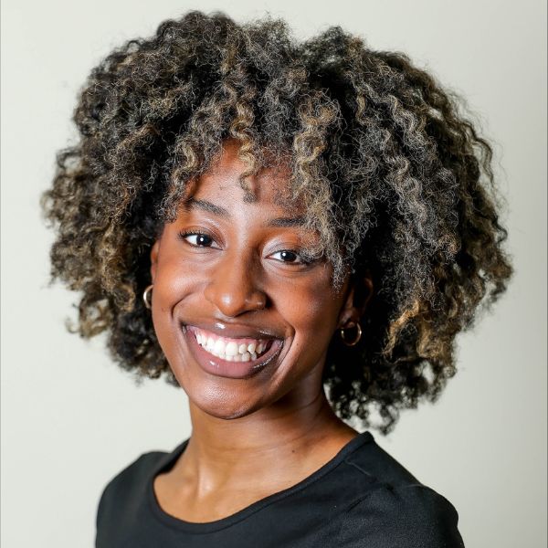 Smiling person with short, curly hair and hoop earrings, wearing a black top, stands against a plain light background.