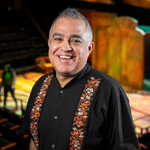 A smiling person with short gray hair stands on a theater stage, wearing a black shirt with orange and white embroidered designs.