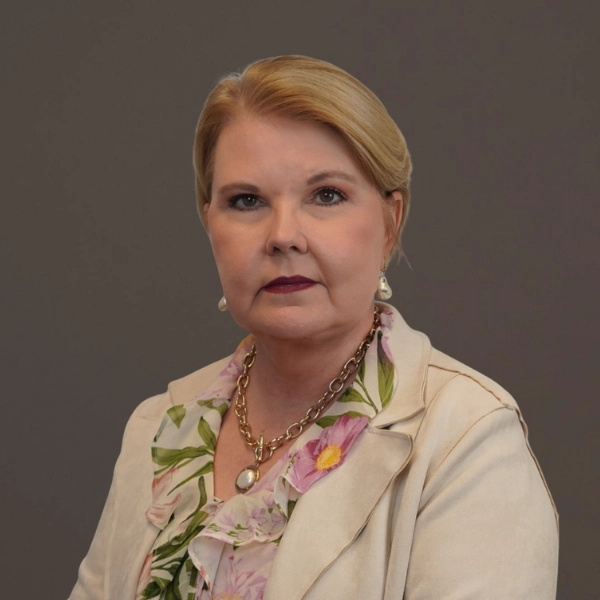 A woman with blonde hair is wearing a light floral blouse, beige blazer, gold chain necklace, and pearl earrings, posing against a plain gray background.