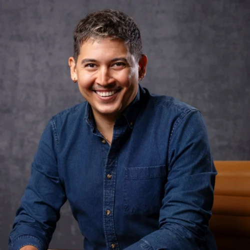 Smiling person with short hair and earrings, wearing a button-up denim shirt, seated in front of a dark mottled background—reminiscent of David Mendizábal’s approachable and creative presence.