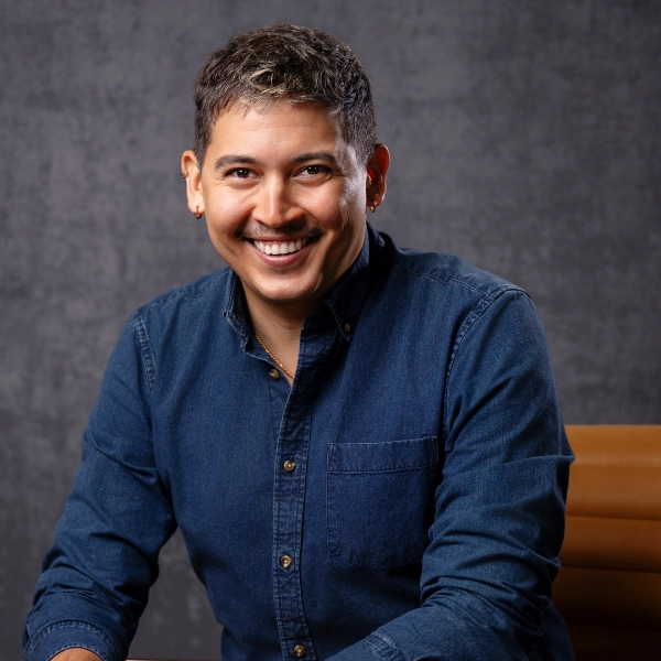 Smiling person with short hair and earrings, wearing a button-up denim shirt, seated in front of a dark mottled background—reminiscent of David Mendizábal’s approachable and creative presence.