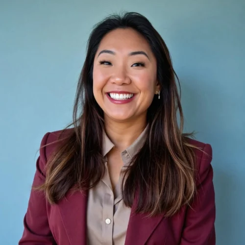 Woman with long dark hair, wearing a maroon blazer and beige blouse, smiles in front of a plain blue background.