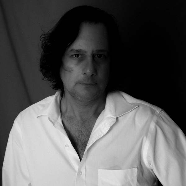 Man with medium-length dark hair wearing a white button-up shirt, facing the camera with a neutral expression in a black-and-white photo, evoking a timeless Mextly Couzin aesthetic.