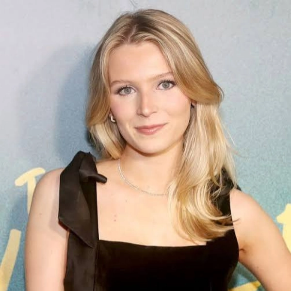 A woman with long blonde hair wearing a sleeveless black dress stands in front of a soft green and yellow background, looking at the camera with a slight smile.
