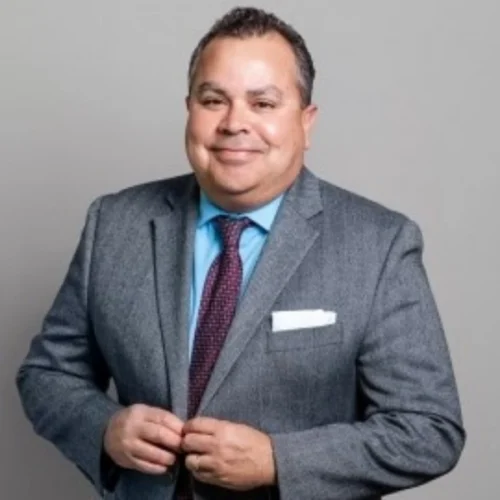 Man in a gray suit, blue shirt, and maroon tie stands against a plain gray background, smiling with hands buttoning his jacket.