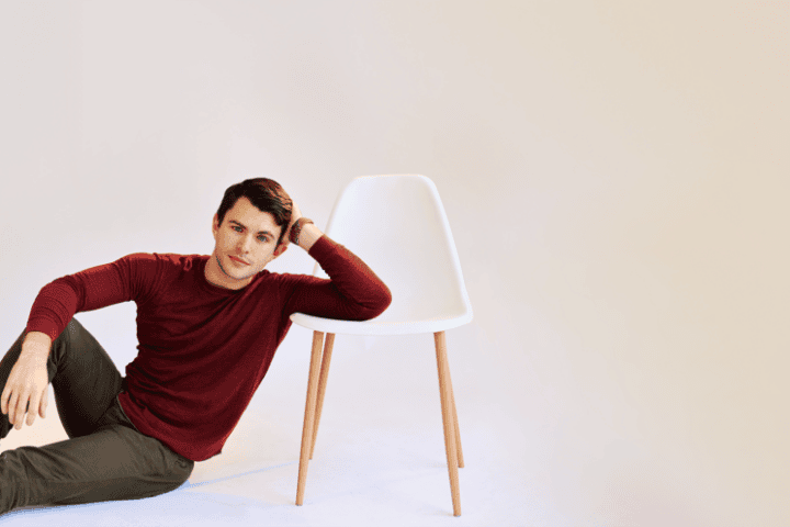 A man in a red shirt sits on the floor, leaning his arm on a white chair with wooden legs, against a plain light background—evoking the quiet introspection found in “A Betrayal of All Betrayals: Jacob Ryan Smith Talks LIKE FATHER.”.