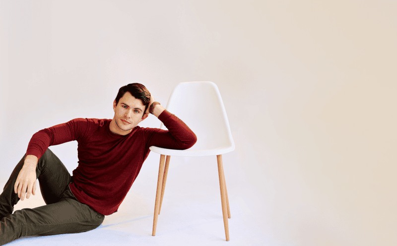 A man in a red shirt sits on the floor, leaning his arm on a white chair with wooden legs, against a plain light background—evoking the quiet introspection found in “A Betrayal of All Betrayals: Jacob Ryan Smith Talks LIKE FATHER.”.