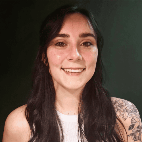 A woman with long dark hair and a floral tattoo on her shoulder smiles at the camera against a dark background.