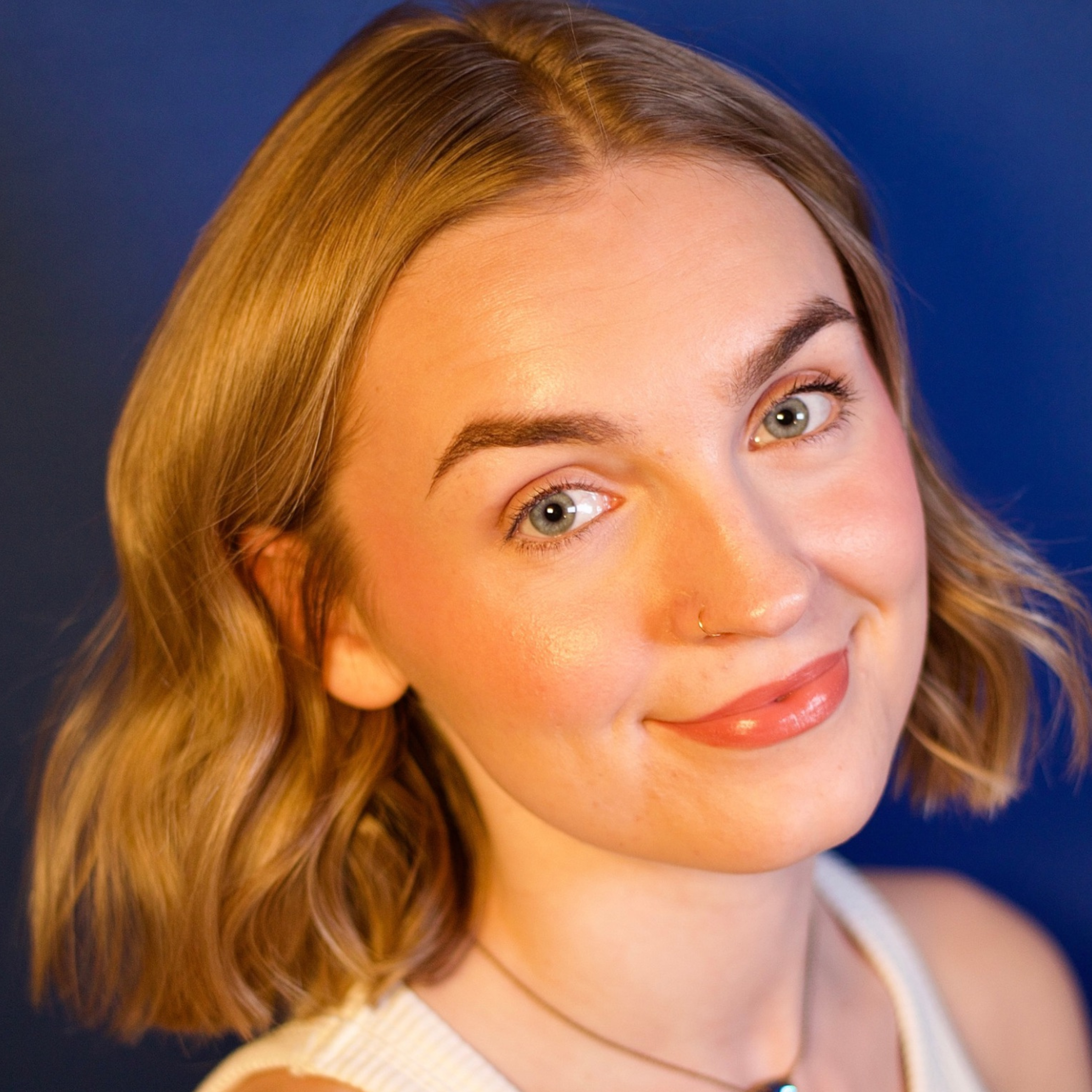 Close-up portrait of a woman with short blonde hair and a nose ring, smiling at the camera against a blue background.