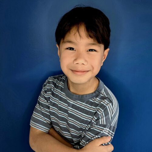 A young boy with dark hair wearing a striped shirt stands with his arms crossed against a blue background.