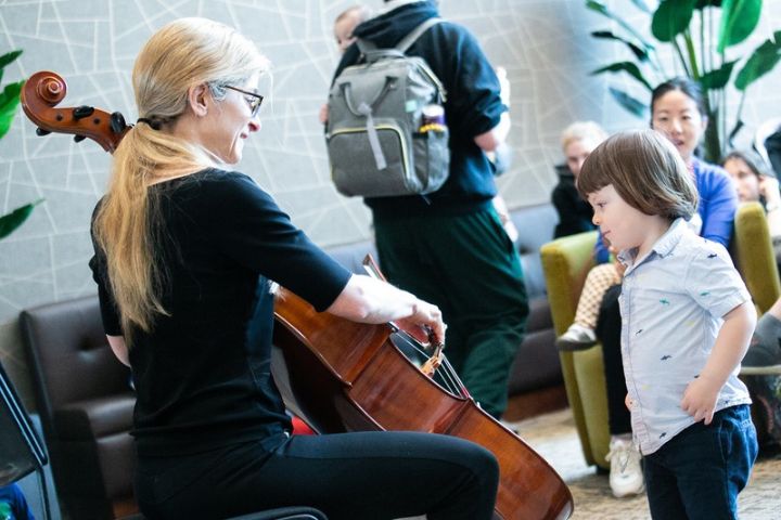 A woman plays the cello for a young child in a PNC PlaySpace room with other people.