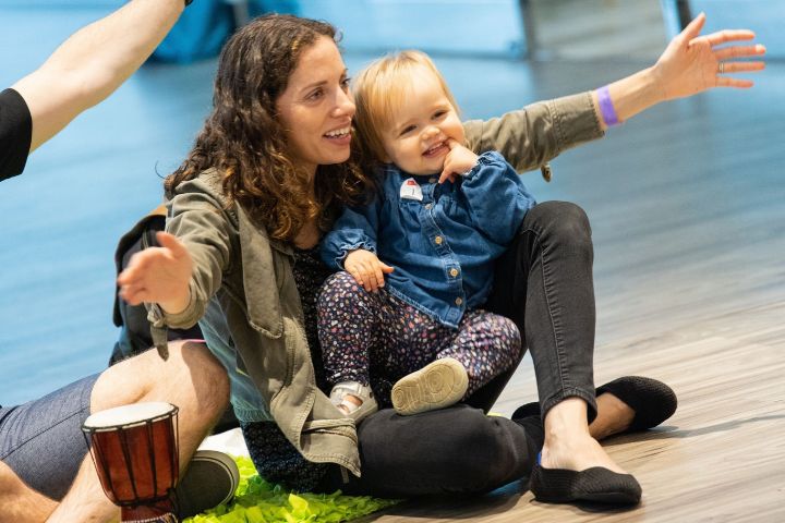A woman sits on the floor in a PNC PlaySpace with a toddler on her lap, both reaching out with their arms.